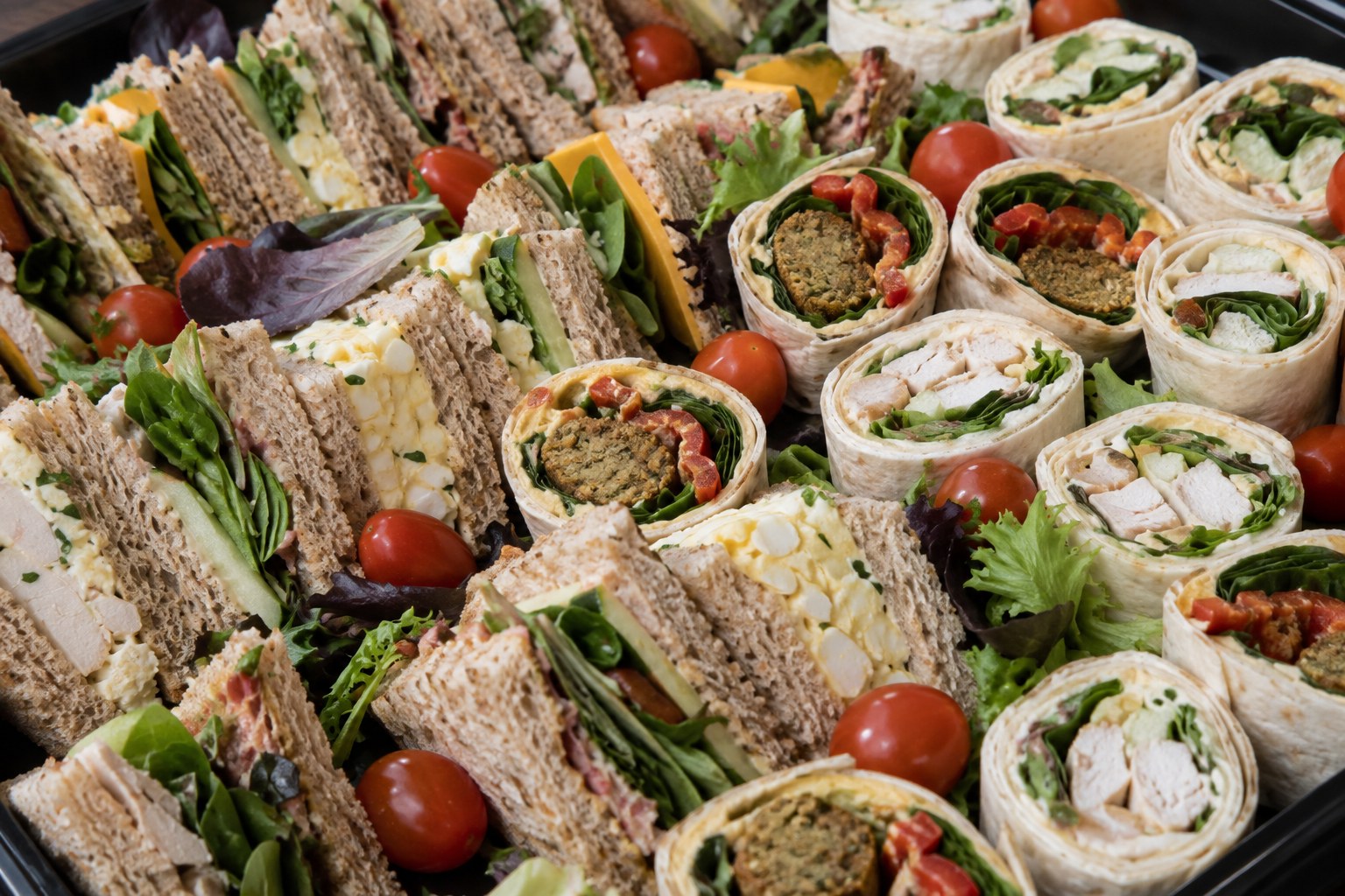 Cut sandwiches and rolled wraps packed in rows with cherry tomatoes and salad leaves — chicken, egg, cheese, and a falafel and roasted pepper wrap