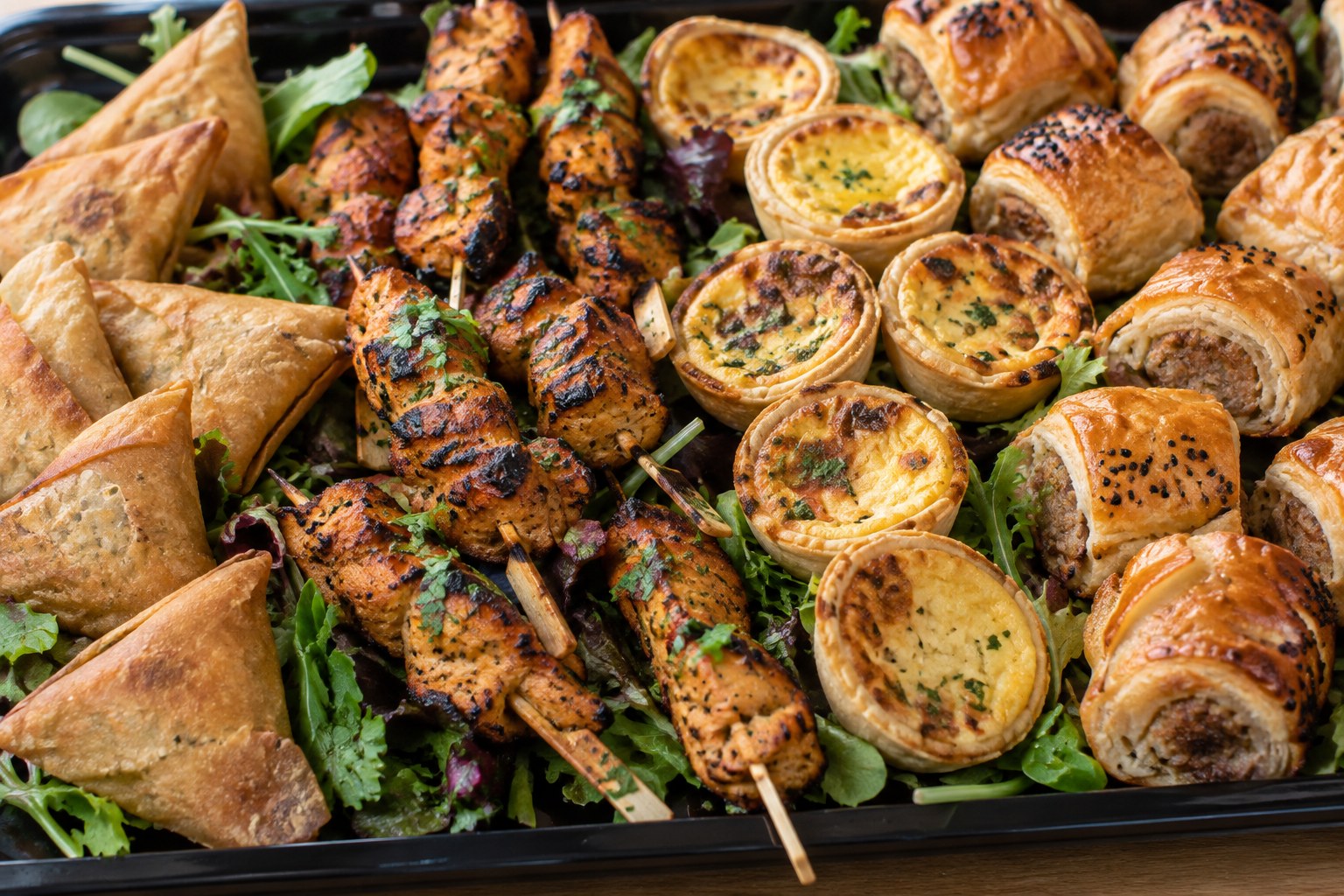 Golden samosas, chicken tikka skewers, mini quiches and mini sausage rolls laid over salad leaves on a black tray